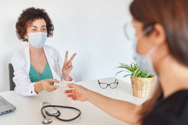Yüz maskeli bir kadın doktor ofisinde bir hastayla ilgileniyor.