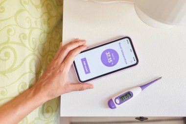 Akıllı telefonu ve bazal termometresi olan bir kadının el görüntüsü.