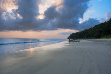 Gün batımında sakin bir tropikal plaj. Dalgalar, dramatik bulutlar ve pastel ışığı yansıtan geniş kumlu bir kıyı şeridi.