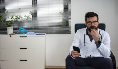 Beyaz önlüklü yakışıklı beyaz erkek doktor enfeksiyoncusunun yakın plan portresi. Klinikteki dolapta oturmuş hasta doktor konseptiyle internet kamerasından görüntülü konuşma yapıyor.