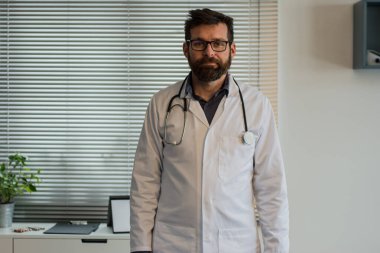 Beyaz önlüklü yakışıklı beyaz erkek doktor enfeksiyoncusunun yakın plan portresi. Klinikteki dolapta oturmuş hasta doktor konseptiyle internet kamerasından görüntülü konuşma yapıyor.