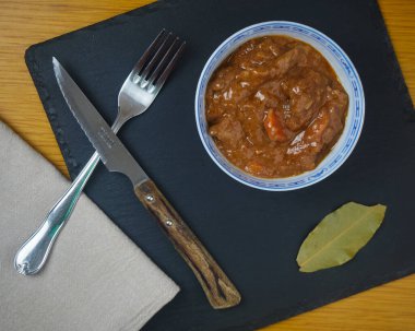 Fricando, geleneksel Katalan tapa, metal çatal bıçakla dekore edilmiş bir kasede sığır güveci, bir peçete ve siyah tahta üzerinde defne yaprağı. Üst görünüm