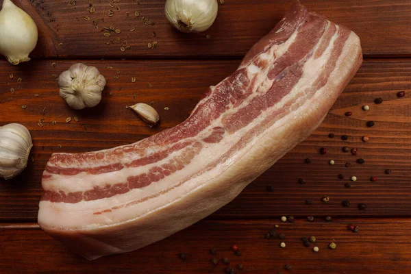 Roher Schweinebauch auf einem Holztisch mit Zwiebeln, Knoblauch und Gewürzen — Stockfoto