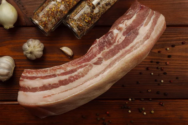 Barriga de porco crua em uma mesa de madeira com cebolas, alho e jarros de temperos — Fotografia de Stock