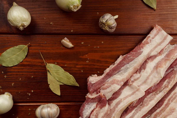 Bacon cru sur une table en bois avec oignon, ail et épices — Photo