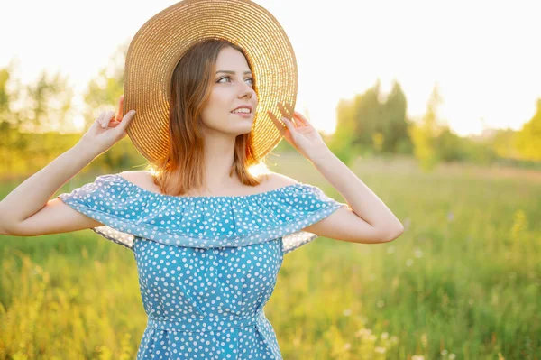 Een jonge vrouw in een veld bij zonsondergang past haar hoed aan en glimlacht — Stockfoto