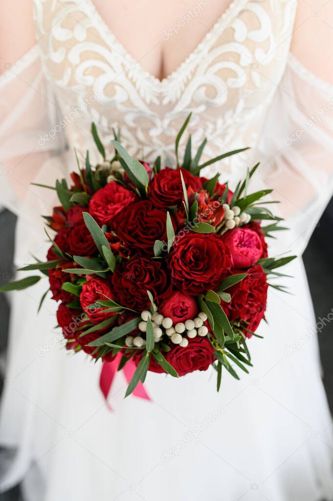 The bride holds a beautiful bouquet of red roses and peonies in her hand.