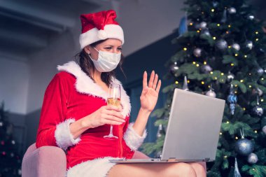 young woman in a Santa Claus costume sits in front of a computer screen and wave his hands. Christmas greetings online. New years in quarantine.