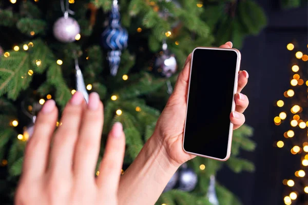 한 여성이 크리스마스 트리를 배경으로 스마트폰 화면에서 인사를 건넵니다. 온라인에서의 인사말의 개념. 새해 코로나 바이러스와 격리 구역이야. 설계를 위한 빈 공간 — 스톡 사진