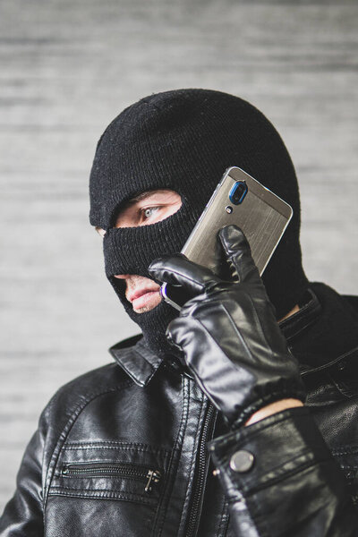 Молодой взрослый в черной одежде со скрытым лицом. Неумышленный мошенник использует мобильный телефон. Мошенник звонит. Мошенничество. Мобильная ракетка. Хакеры угоняют по телефону. Мошенничество на мобильных телефонах.