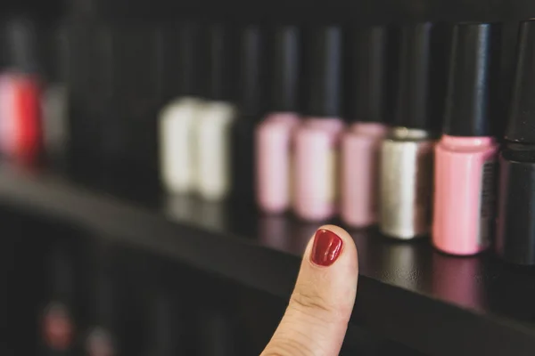 The choice of nail polish. A finger with a red-painted fingernail points at the bottles of nail polish. lots of nail polish options on the shelf in the store.