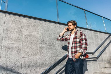 Telefonla konuşan modern yakışıklı genç adamın portresi. Elinde cep telefonu olan hippi bir adam. Şehir merdivenleri, şehir sokağı.