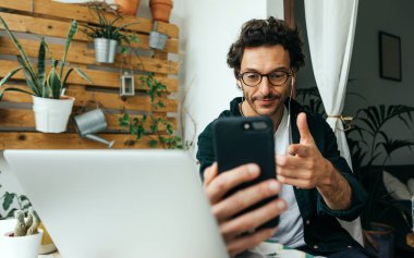 Bilgisayarlı ve akıllı telefonlu yakışıklı adam evden uzaktan çalışıyor, cep telefonuyla video görüşmesi yapıyor. Uzaktan kumandada serbest çalışan biri. Rahat iş yeri