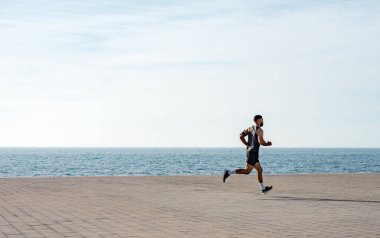 Genç ve zinde bir adam deniz kenarında koşuyor. Erkek koşucu sabah erkenden koşuyor. Sporcu dışarıda spor yapıyor. Spor salonunun dışındaki kilitlenme sırasında egzersiz..
