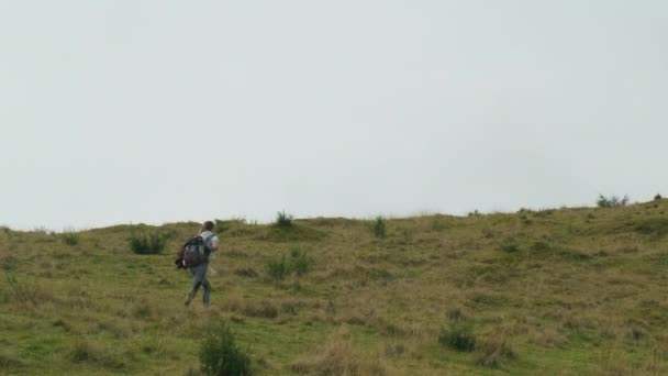Mooi meisje met een rugzak loopt door het gras op een groene heuvel in de middag. grijze lucht, wijd beeld — Stockvideo
