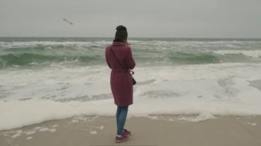 Marsal kürklü bir kız, soğuk kış denizinin arka planında, gri gökyüzünde dalgalar ve sisli havayla uçan martılara bakar.