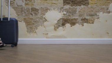 Bir kız tatile çıkıyor, mavi bir bavulu önüne yuvarlıyor mavi bir elbise ve siyah topuklu sandaletler giyiyor, kapalı bir yerde. Yan görünüm, yakın çekim, yavaş çekim