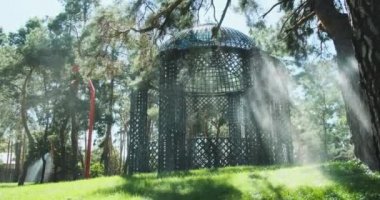 Güneşli bir günde yeşil çimlerin üzerinde parktaki çardak. Çam ağaçları arasında fıskiyeler, su damlaları düşüyor, orta düzey planlar, yavaş çekimde.