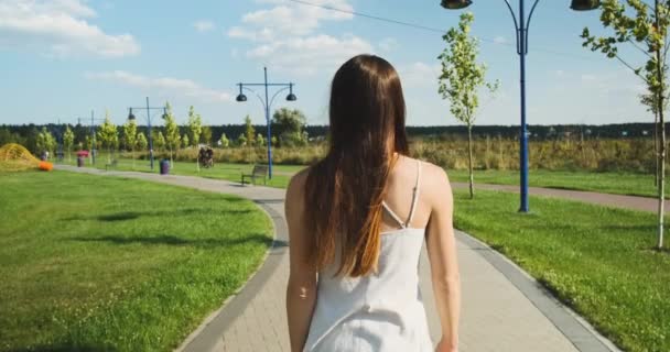 Jong meisje in een witte jurk loopt in het park op een heldere dag, een pad gemaakt van tegels, bomen, achteraanzicht, in slow motion — Stockvideo