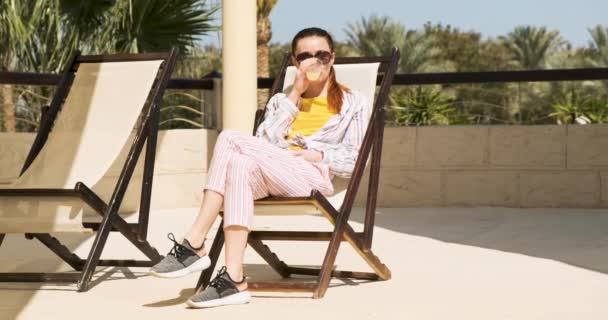 Una chica se sienta en un chaise lounge y bebe una copa de una copa, disfrutando del descanso. Día despejado, cielo azul, palmeras, imágenes generales, cámara lenta — Vídeos de Stock