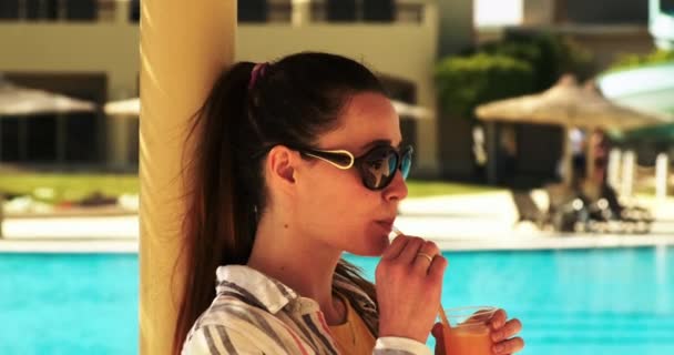 Gros plan, une fille solitaire aux cheveux roux en lunettes de soleil boit un cocktail dans une tasse en plastique dans un complexe près de la piscine, au ralenti — Video
