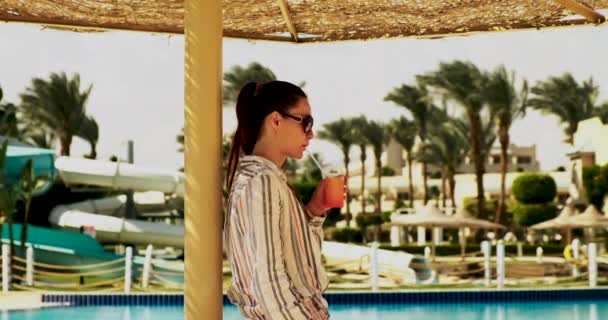 Mooie jonge vrouw, in het resort onder een paraplu bij het zwembad, een cocktail drinken en genieten van de rest. Heldere lucht, zon, medium beelden — Stockvideo