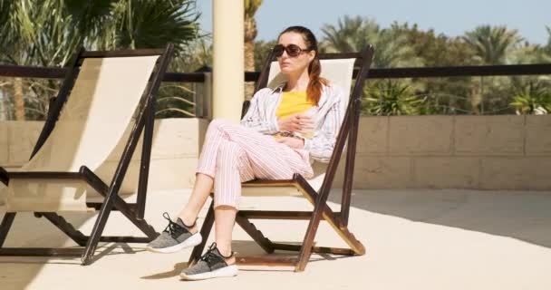 Beautiful brown-haired woman sitting on a sun lounger at the resort and drinking a cool drink. Clear day, brighter sun, palm trees. — Stock Video