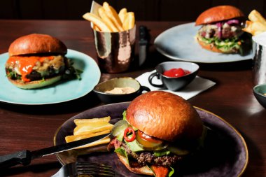 Different burgers on the table, served frenc fries and sauces on plates. The closest spicy beef burger with jalapenos, pickled cucumbers and avocado with spicy sauce on a plate with fork and knife.