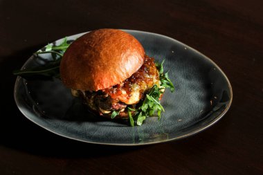 Ev yapımı ekmeğin üzerine karamelli soğan, peynir ve roka ile sulu sığır etli burger. Hamburger, koyu kahverengi masadaki el yapımı gri tabakta servis ediliyordu. Koyu arkaplan, yakın plan, üst görünüm.