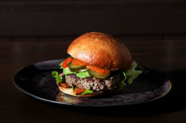 Juicy spicy beef burger with slices of pickled cucumbers, avocado, hot sauce, piece of red pepper and lettuce on a homemade bun on a handmade plate on a dark brown table. Dark background, dark plate.