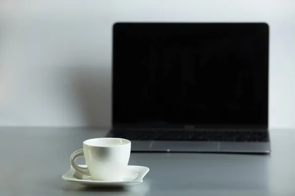 Composition with a modern opened laptop a white coffee cup and saucer on the table on a light gray background. Remote workplace, work from home. Moke up with laptop and coffe cup.