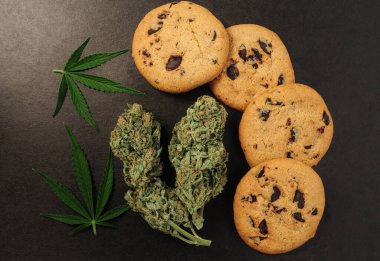 CBD yağlı tatlılar. Siyah arka planda marihuana tomurcukları. Kenevir çiçekleri ve fındıklı çikolatalı kurabiyeler. Üst manzara, düz uzanma.