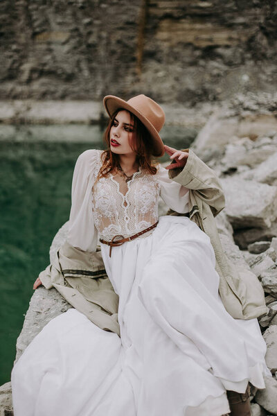 fashionable woman in a white dress sitting on rock at lake 