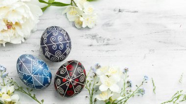 Three handmade painted Easter eggs with spring flowers. Ukrainian pysanka decorated with wax-resist dyeing technique on white shabby wooden background with empty space for text clipart