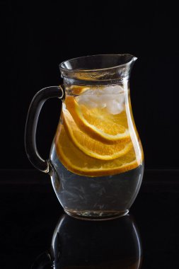 Su, dilimlenmiş portakal ve siyah arka plan üzerine buz ile sürahi