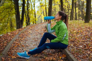 Koşucu sonbahar parkında antrenmandan sonra dinleniyor. Yolda oturan su içen bir kadın. Sportif aktif yaşam tarzı. Temiz havada sonbahar egzersizi.