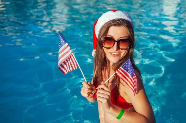 Yeni yıl ve Noel kutlaması. Noel Baba şapkalı ve bikinili bir kadın yüzme havuzunda ABD bayrakları tutuyor. Tropik kış tatili