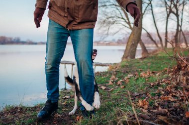 Sonbahar parkında göl kenarında köpekle oynayan adam. Mutlu ev hayvanı sahibinin bacağına sarılırken dışarıda gezerken eğleniyor. Sonbahar etkinlikleri