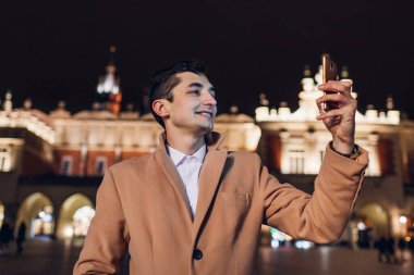 Turist, Krakow Polonya 'daki pazar meydanında gece yürürken akıllı telefondan selfie çekiyor. Sonbaharda Avrupa seyahati