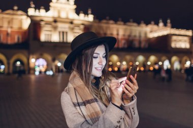 Krakow Polonya 'nın pazar meydanında gece akıllı telefon kullanan turist kadın. Kız şehir merkezinde sosyal medyayı kontrol ediyor. Avrupa 'yı gezmek