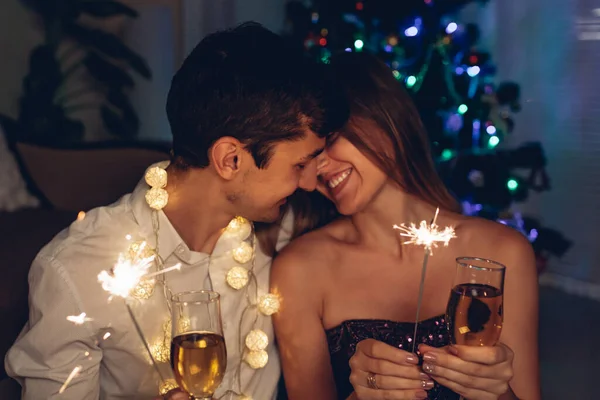 Christmas and New year party. Couple in love burning sparklers by illuminated Christmas tree with champagne at home. Celebrating winter holidays