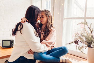 Anne ve kızı dekore edilmiş Noel ağacının yanında mutfak penceresinin eşiğinde sarılıyorlar. Aile evde iyi vakit geçiriyor.