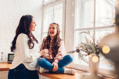 Anne ve kızı dekore edilmiş Noel ağacının pencere eşiğinde dinleniyorlar. Aile evde birlikte gülerek eğleniyor. Tatil kutlamalarının tadını çıkarıyor.