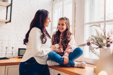 Anne ve kızı dekore edilmiş Noel ağacının pencere eşiğinde dinleniyorlar. Aile evde birlikte gülerek eğleniyor. Tatil kutlamalarının tadını çıkarıyor.