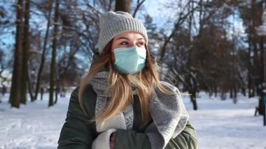 Kar parkında yürüyen koruyucu yüz maskesi takan bir kadının kış portresi. Corona virüsü covid-19 salgın güvenlik önlemleri.
