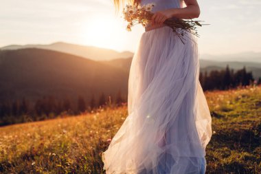 Gelin, gün batımında dağlarda elinde buket tutan mavi bir gelinlik giyiyor. Papatya çiçekleri içinde çayırda yürüyen bir kadın
