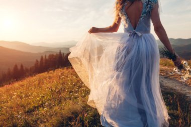 Gelin, gün batımında dağlarda elinde buket tutan mavi bir gelinlik giyiyor. Papatya çiçekleri içinde çayırda yürüyen bir kadın