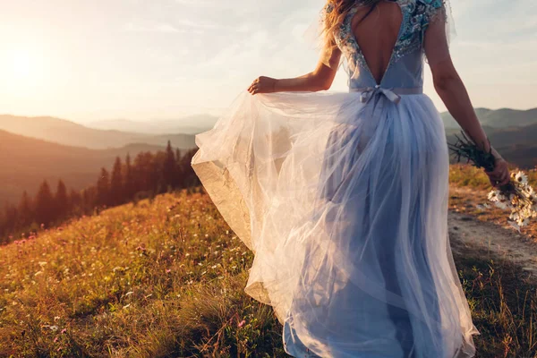 Gelin, gün batımında dağlarda elinde buket tutan mavi bir gelinlik giyiyor. Papatya çiçekleri içinde çayırda yürüyen bir kadın