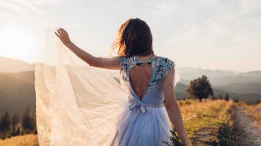 Güzel gelin, gün batımında dağlarda, üzerinde nakışla mavi bir gelinlikle dans ediyor. Kadın çiçekleri tutuyor. Karpatlar 'da Düğün