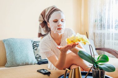 Kadın banyodan sonra evde orkide çiçeklerinin keyfini çıkarırken yüz maskesi takmış. Doğal kozmetik ürünleri. Cilt bakımı
